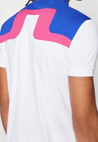 Vit polotröja med blå och rosa detaljer vid kragen och axelområdet, med en slät yta och korta ärmar.