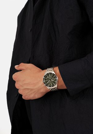 Sølv rustfrit stål ur med en rund sort chronograph urskive, teksturerede detaljer og et sammensat metalarmbånd, båret på et håndled.