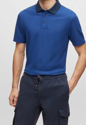 Blauwe polo met een gestructureerde afwerking, korte mouwen en een marineblauwe kraag. Bevat drie zwarte knopen aan de voorkant. Gecombineerd met donkere cargobroeken.