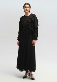 Juoda suknelė, turinti sluoksniuotą, raukšlėtą audinį ant rankovių ir viršutinės dalies, su laisvai krintančia sijona. Pagaminta iš lengvos medžiagos, kulkšnies ilgio.