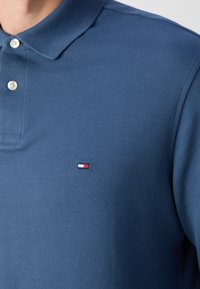 Granatowa koszulka polo z fakturowanej tkaniny, z dwiema guzikami przy dekolcie i małym wyszywanym logo w kolorze czerwonym, białym i niebieskim.