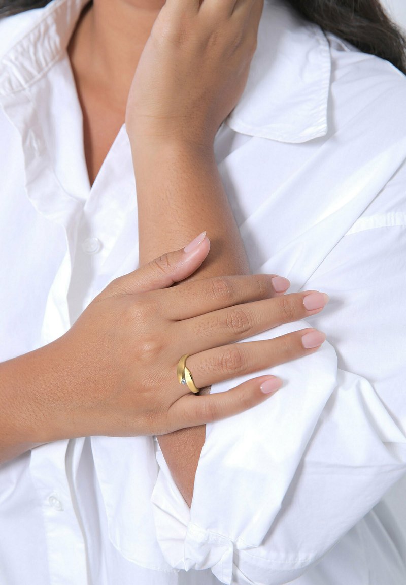 Goldring mit einem Diamantakzent, getragen an einer Hand mit einer hellrosa Maniküre, vor einem weißen Shirt mit hochgekrempelten Ärmeln. Glatte Hauttextur.