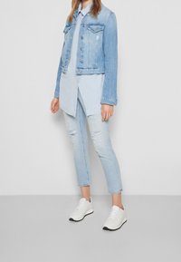Chaqueta de denim azul claro con cuello puntiagudo y herrajes plateados, llevada sobre una camisa azul claro y unos jeans ajustados de lavado claro desgastado. Zapatillas blancas.