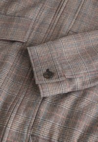 Tessuto a quadretti con una palette di colori marrone e grigio, caratterizzato da sottili linee rosse e blu. Include un bottone nero su un polsino.