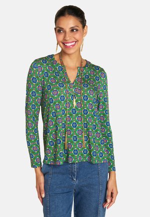 Grün gemusterte Bluse mit langen Ärmeln, die florale Motive in Blau und Pink zeigt. Bemerkenswerte Details sind ein V-Ausschnitt und eine Halskette mit goldfarbener Anhänger.