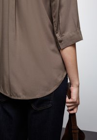 Blusa marrone leggera con una texture liscia, maniche arrotolate 3/4, orlo arrotondato, abbinata a jeans scuri; mano che tiene una tracolla.