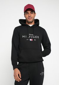 Zwarte hoodie met buidelzak aan de voorkant, met witte tekst "Tommy HI-FIGER 1985" en een klein logo. Gecombineerd met zwarte joggingbroek.