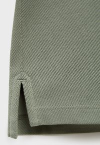 Tessuto di cotone verde con una maglia testurizzata, caratterizzato da un orlo ventilato per la traspirabilità e dettagli cuciti lungo i bordi.