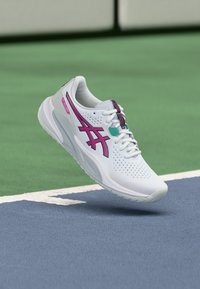 Valge sportlik jalanõu perforatsiooniga tekstuuriga, roosade ja türkiissiniste detailidega ning sportliku disainiga, eksponeeritud rohelise väljakutaustaga.