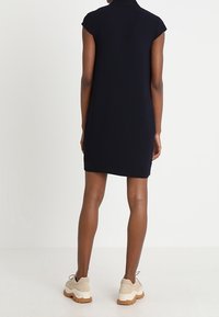 Woman standing facing away wearing a short-sleeve black dress and beige chunky sneakers against white background.