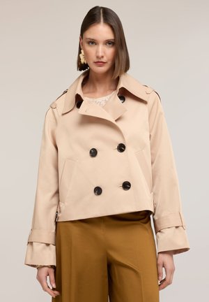 Vrouw die een beige dubbelgeknoopte cropped trenchcoat met zwarte knopen en mosterdkleurige wijde broek draagt, staand tegen een effen achtergrond.