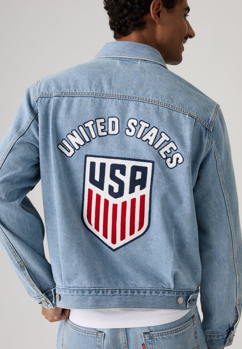 Homme portant une veste en jean bleu clair avec "United States" et un logo de bouclier des États-Unis en rouge, blanc et bleu dans le dos.