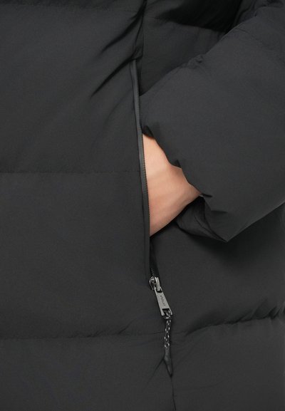 Ročna vstavljen v stranski žep z zadrgo črne podložene jakne z vidnim držalom za zadrgo in detajli šivanja.