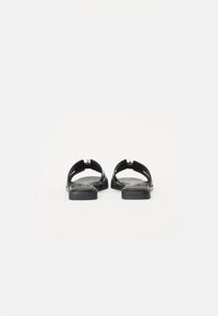 Sorte sandaler med flat såle, åpen bak og utskjæringsdesign i remmene. Har en liten gullakcent på hælen.