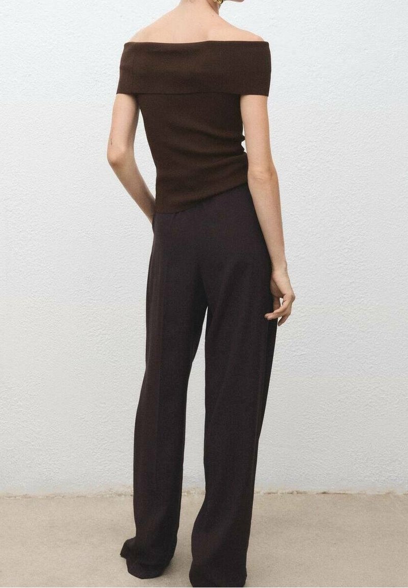 Femme debout, de dos, portant un haut marron foncé à épaules dénudées et un pantalon noir à jambes larges, devant un mur blanc uni.