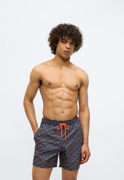 Bărbat tânăr cu păr creț, purtând șorturi de baie bleumarin cu mici modele florale colorate și un șnur portocalie, pozând pe un fundal alb.