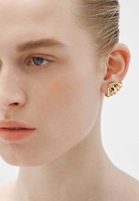 Pendientes de metal dorado con un diseño texturizado en forma de abanico. Presentan superficies lisas y un acabado pulido, contrastando con el tono de piel.