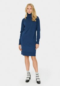 Mörkblå turtleneckklänning med långa ärmar, ribbade manschetter och rak form. Bärs med grå strumpor och svarta klackskor.