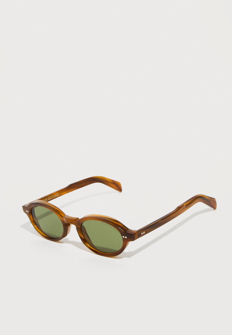 Lunettes de soleil ovales en écaille de tortue marron avec des verres verts posées sur une surface blanche.