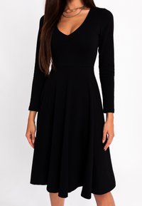 Femme portant une robe midi noire à manches longues et décolleté en V, avec une taille cintrée et une jupe évasée, accessoirisée d'un collier à chaîne fine.