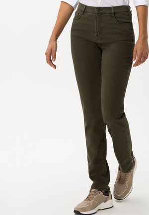 Person wearing olive green pants, beige sneakers, and a white shirt, walking against a plain white background.