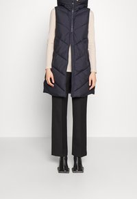 Chaleco acolchado azul marino con cremallera frontal, patrón de rombos en chevron y capucha. Combinado con pantalones anchos negros y botas hasta el tobillo. Top de manga larga de color claro.