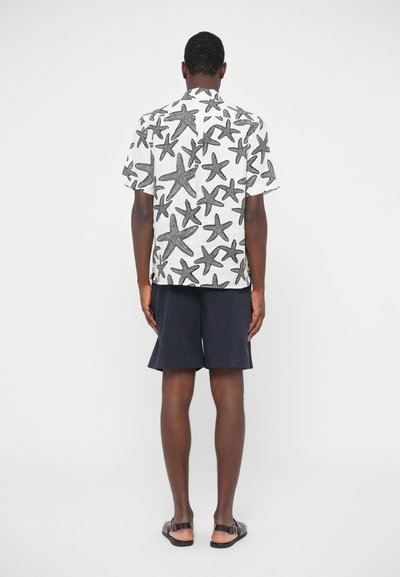 Kratki rokavicasti srajč z belim ozadjem in črnimi morskim prstom, v kombinaciji s temno modrimi kratkimi hlačami in rjavimi sandali, pogled zadaj.