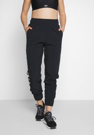 Femme portant un pantalon de jogging noir à bords élastiques, poches latérales, et des chaussures de sport noires avec des lacets à motifs, debout devant un fond blanc.