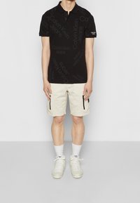 Man som bär en svart Calvin Klein Jeans polo, beige cargoshorts med svarta dragkedjor, vita strumpor och vita sneakers, står mot en vit bakgrund.