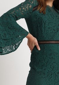 Žena v zelených krajkových šatech s volnými rukávy a síťovaným pasem, s rukou odpočívající na boku.