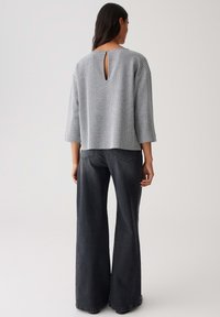 Femme de dos portant un haut gris à motifs avec une fente dans le dos et un jeans noir à jambes larges, se tenant devant un fond uni.