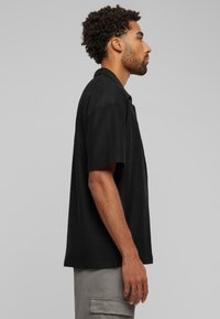 Black short-sleeve shirt in a textured fabric, featuring a collar and a straight cut, displayed on a model from a side angle against a gray background.