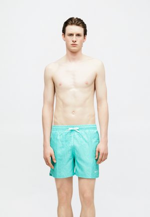 Νεαρός άνδρας που στέκεται μπροστά, φοράει τυρκουάζ μαγιό με λευκό κορδόνι και διακριτικό σχέδιο κυμάτων πάνω σε απλό λευκό φόντο.