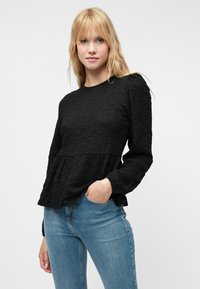 Czarna, fakturowana bluzka z długim rękawem, z gromadzonym w talii krojem, z okrągłym dekoltem i subtelnym wzorem. Połączona z jasnoniebieskimi, dopasowanymi dżinsami.