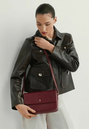 Kobieta ubrana w czarną skórzaną kurtkę, białe spodnie, trzyma bordową skórzaną torebkę na ramię z klapą i złotym zapięciem.