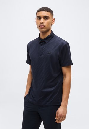 Lühikeste juustega mees, kes kannab tumeda mereväe sinist polo särki väikese valge logoga rinnal, seisab käed taskutes.