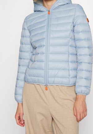 Jasnoniebieska pikowana kurtka z kapturem i przednim zamkiem, noszona z beżowymi spodniami, częściowo widoczny tors i ramiona.