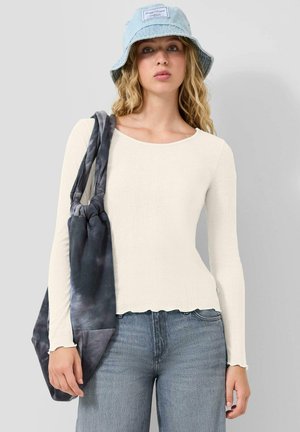 Junge Frau trägt einen hellblauen Anglerhut, ein cremefarbenes, geripptes Langarmshirt, blaue Jeans und trägt eine dunkle Batik-Tasche über der Schulter.