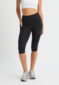 Leggings capri negros hechos de tela elástica que cuentan con una cintura alta y bolsillos laterales, complementados con un logo blanco en la parte inferior de la pierna.