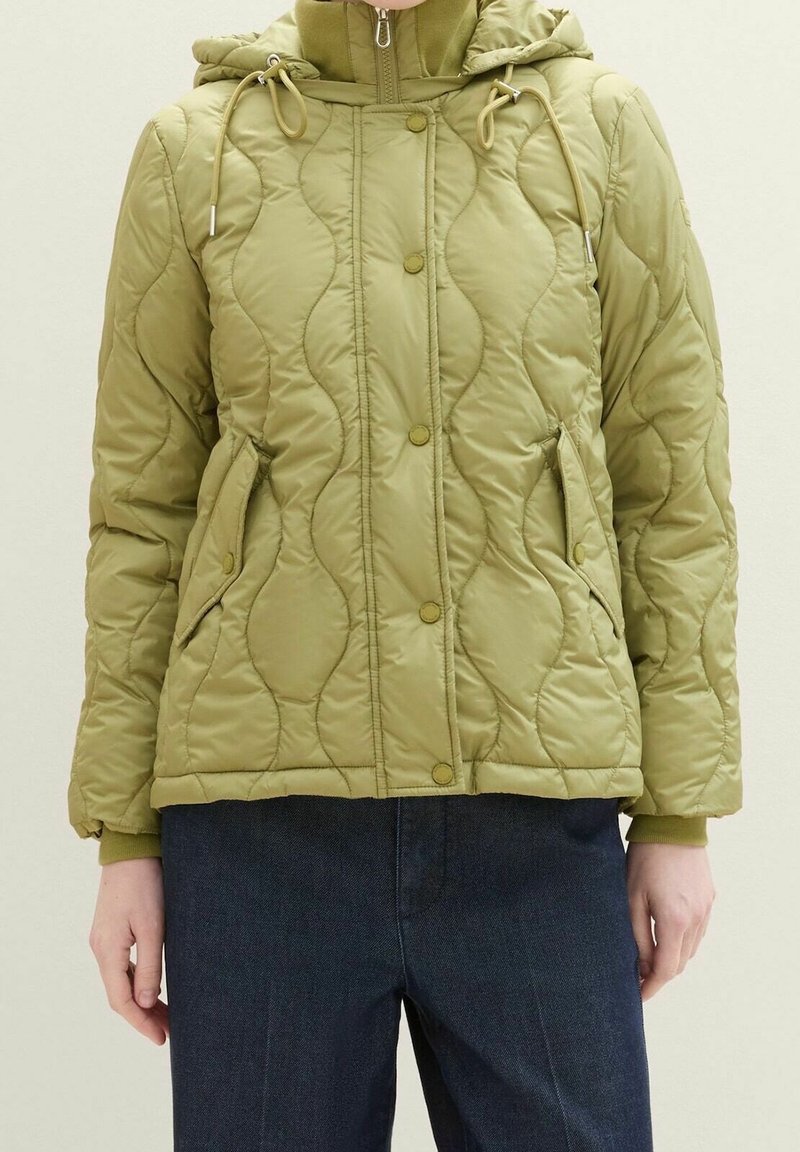 Veste matelassée vert olive avec boutons-pression à l'avant, fermeture éclair, deux poches à boutons-pression et une capuche ajustable portée avec un pantalon foncé.
