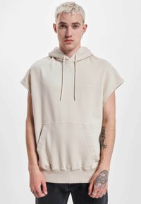 Beige ärmellose Kapuzenjacke aus Baumwollmischung, mit einer Kängurutasche, verstellbarem Kapuzenzug und geripptem Saum. Textlogo auf der Brust.