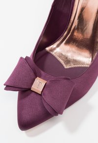 Gros plan sur une chaussure violette en satin à bout pointu avec un nœud assorti et un accent carré doré gravé de la lettre "T" à l'avant.
