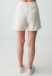 Shorts en lin beige avec une texture lisse, dotés d'une poche arrière et d'un ourlet roulé, associés à des baskets blanches.