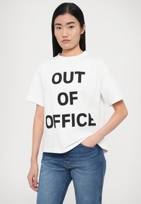 Λευκό βαμβακερό T-shirt με έντονο μαύρο κείμενο "OUT OF OFFICE." Κυβικό σχήμα με κοντά μανίκια και στρογγυλή λαιμόκοψη, συνδυασμένο με τζιν παντελόνι.