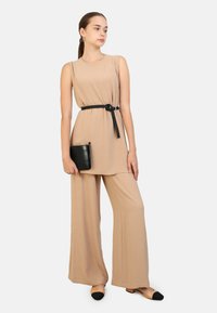 Beige mouwloze top met zijknopen, wijde broek en zwarte riem. Een zwarte clutch en schoenen met zwarte teenaccenten maken de look compleet.