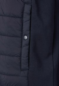 Marineblaue Jacke mit einer gesteppten Textur auf einer Seite, glattem Stoff auf der anderen Seite und einem Druckknopfdetail in der Nähe der Naht.