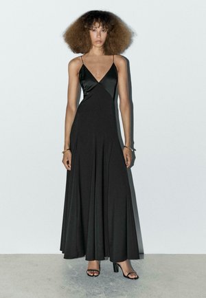 Robe maxi noire avec un décolleté en V profond et des fines bretelles, faite d'un tissu lisse. Elle a une jupe évasée et des plis subtils, associée à des talons noirs.