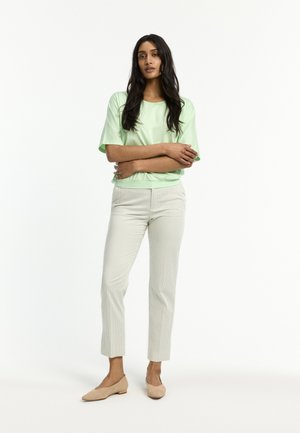 Woman with long dark hair standing with arms crossed, wearing a light green short-sleeve top, light beige pants, and beige flats.