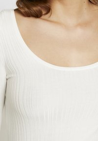 Haut blanc à manches longues côtelées avec un large décolleté en forme de goutte, présentant une texture lisse et un éclat subtil.