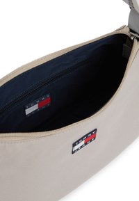 Μπεζ υφασμάτινη τσάντα ώμου με φερμουάρ, με εσωτερικό σε ναυτικό μπλε και ετικέτα με το λογότυπο "TOMMY JEANS" σε κόκκινο, λευκό και μπλε.